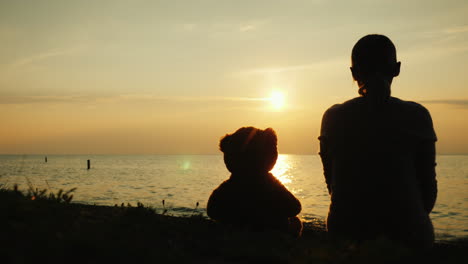 silhouette of a woman and a teddy bear on the lake looking forward over the horizon where the sun se