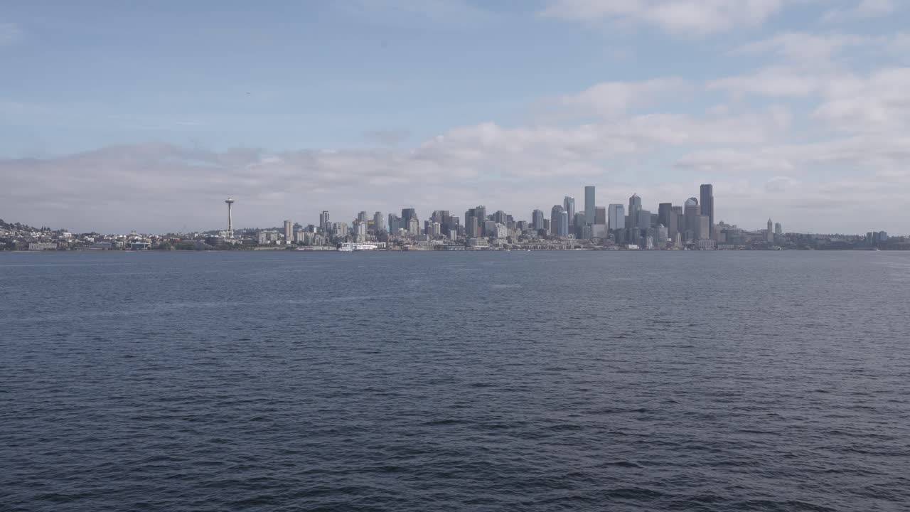 A ferry glides across Puget Sound towards Seattle, offering a wide view of the city’s skyline, with the tall skyscrapers under a partly cloudy sky.