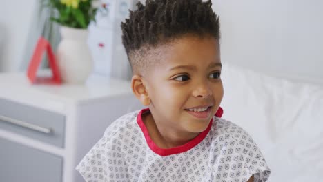 Happy-african-american-boy-patient-sitting-up-in-bed-at-hospital,-smiling