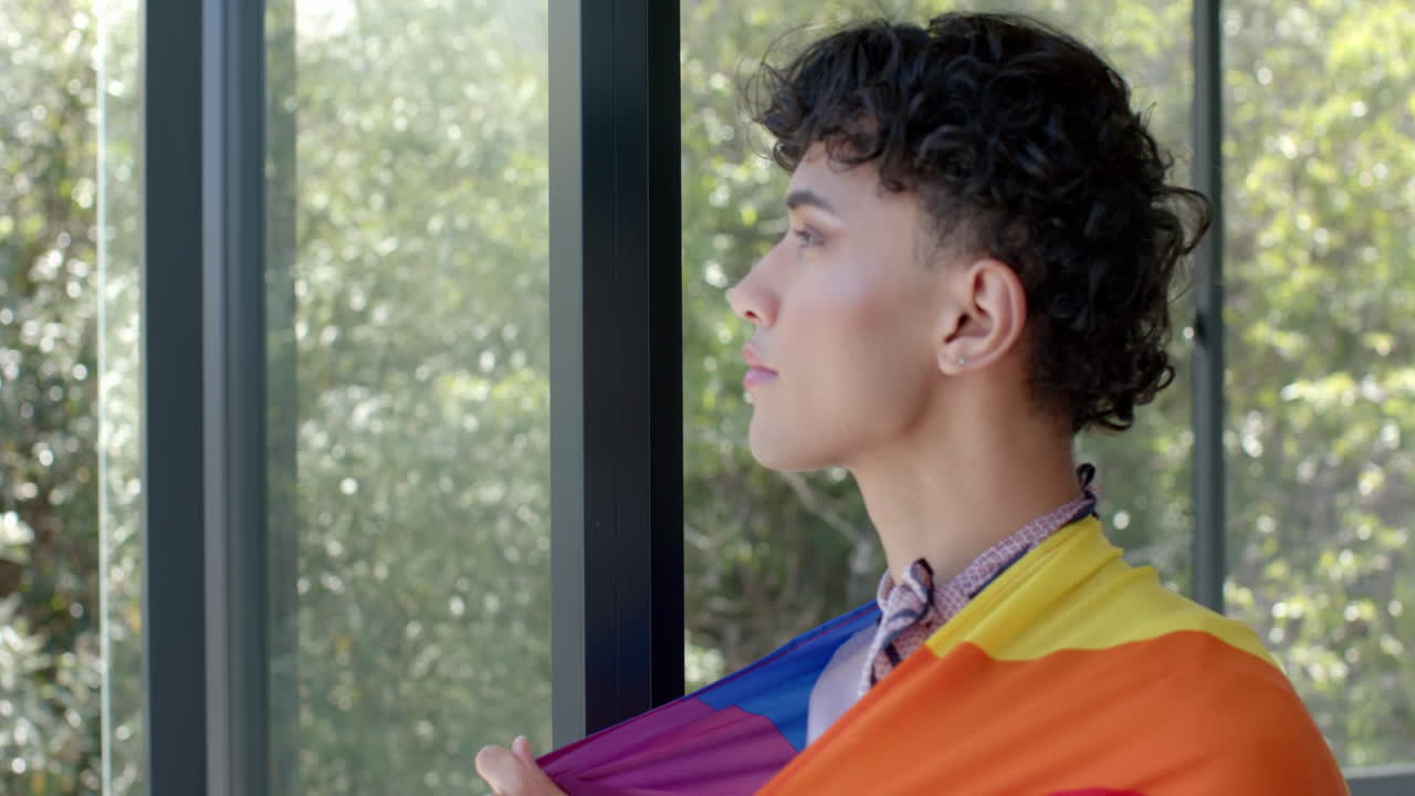 Wrapped in pride flag, person standing by window, looking outside thoughtfully