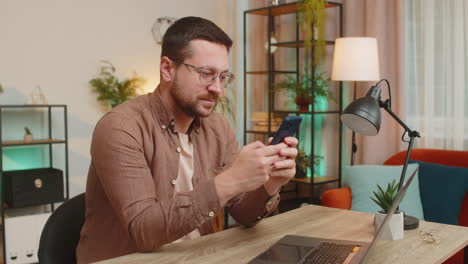 happy young male freelancer sitting at desk using smartphone texting smiling at home apartment