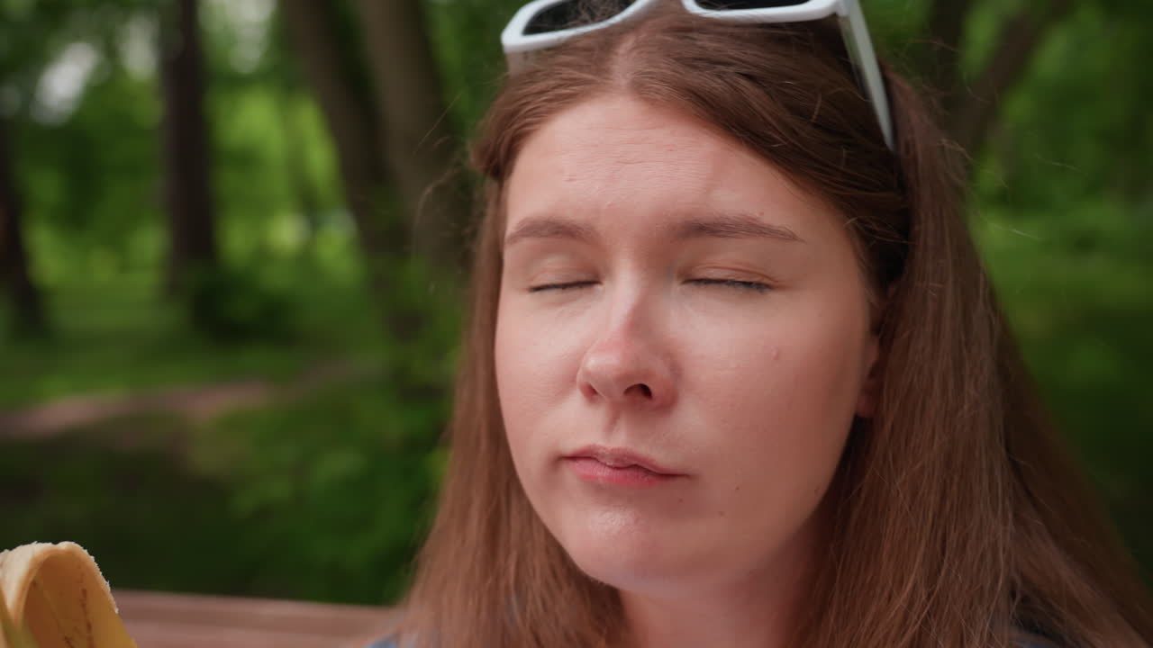 señora elegante saboreando plátano crudo mientras está sentada en un banco de madera en un exuberante parque forestal, mirando de lado con expresión curiosa, disfrutando de un momento de tranquilidad