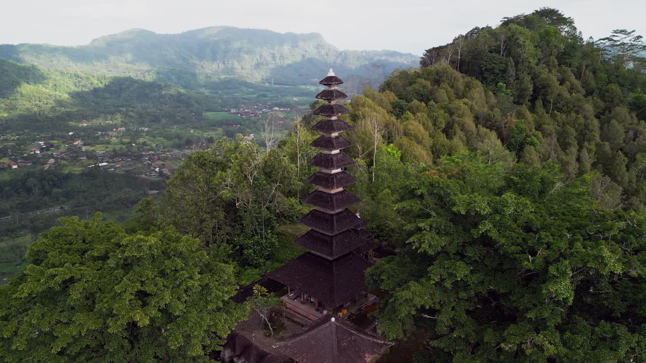 Aerial footage of a Balinese temple complex in Karangasem, surrounded by forested hills and distant mountains. The calm weather and detailed temple rooftops show Bali’s rich cultural landscape
