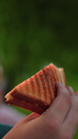 vista parcial de cerca de una persona sosteniendo un sándwich a la parrilla con un fondo ligeramente borroso de personas comiendo, al aire libre, entorno natural con iluminación suave