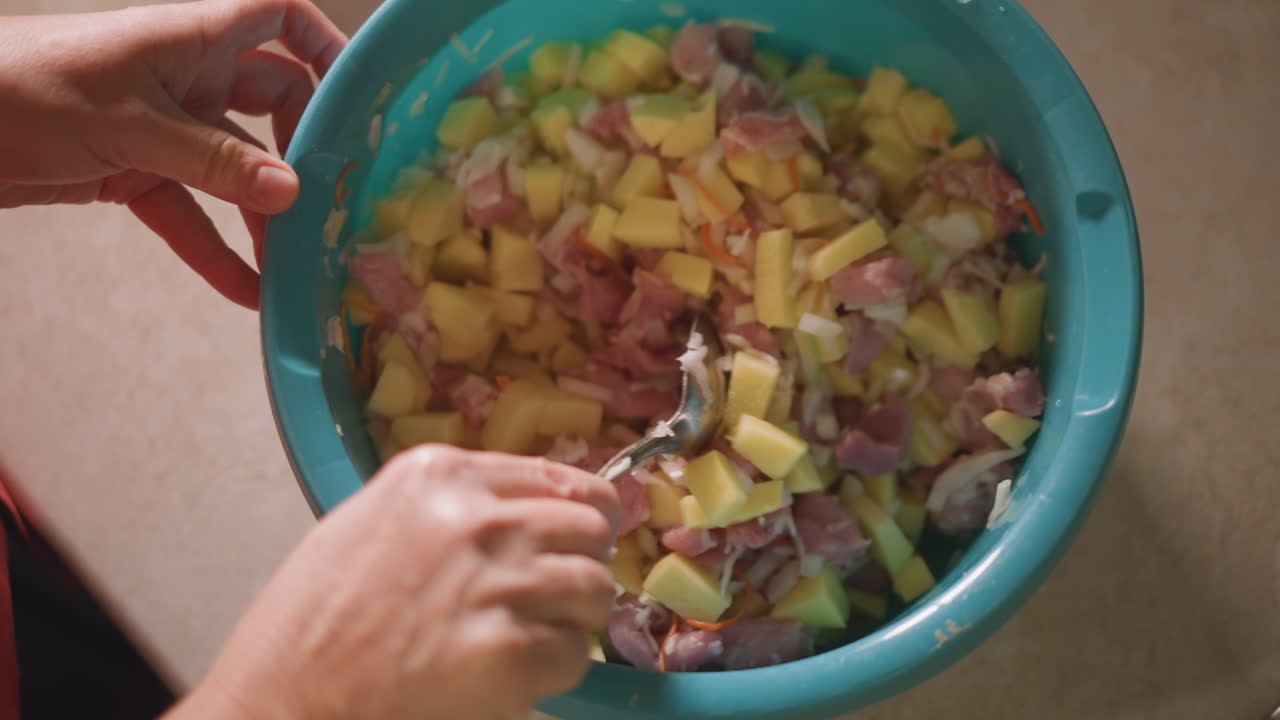 Vista cenital de ensalada de patata y jamón en un cuenco azul, cocinero casero mezclando metódicamente ingredientes picados con cuchara, preparación en encimera para reunión familiar, luz cálida y ambiente rústico casero, listo para servir.