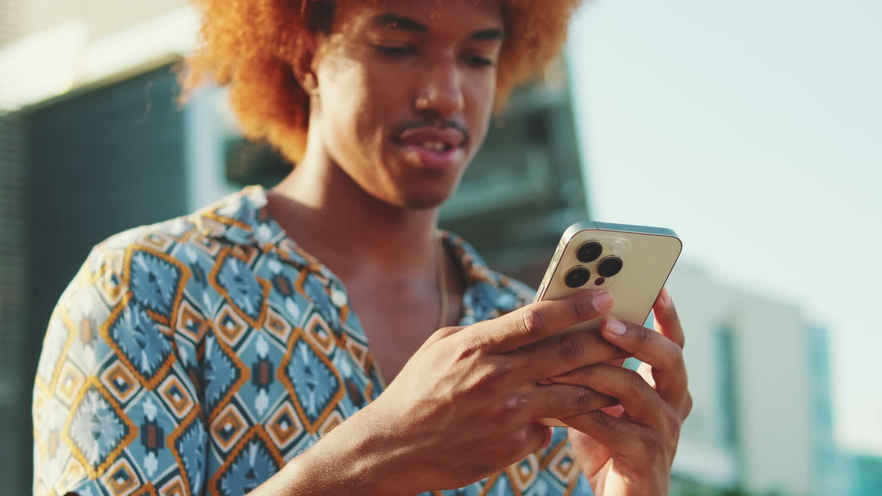 Mladý muž s kudrnatými vlasy se usmívá při používání smartphonu venku