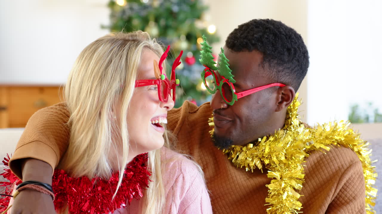 diverse couple wearing festive glasses and tinsel, smiling by Christmas tree