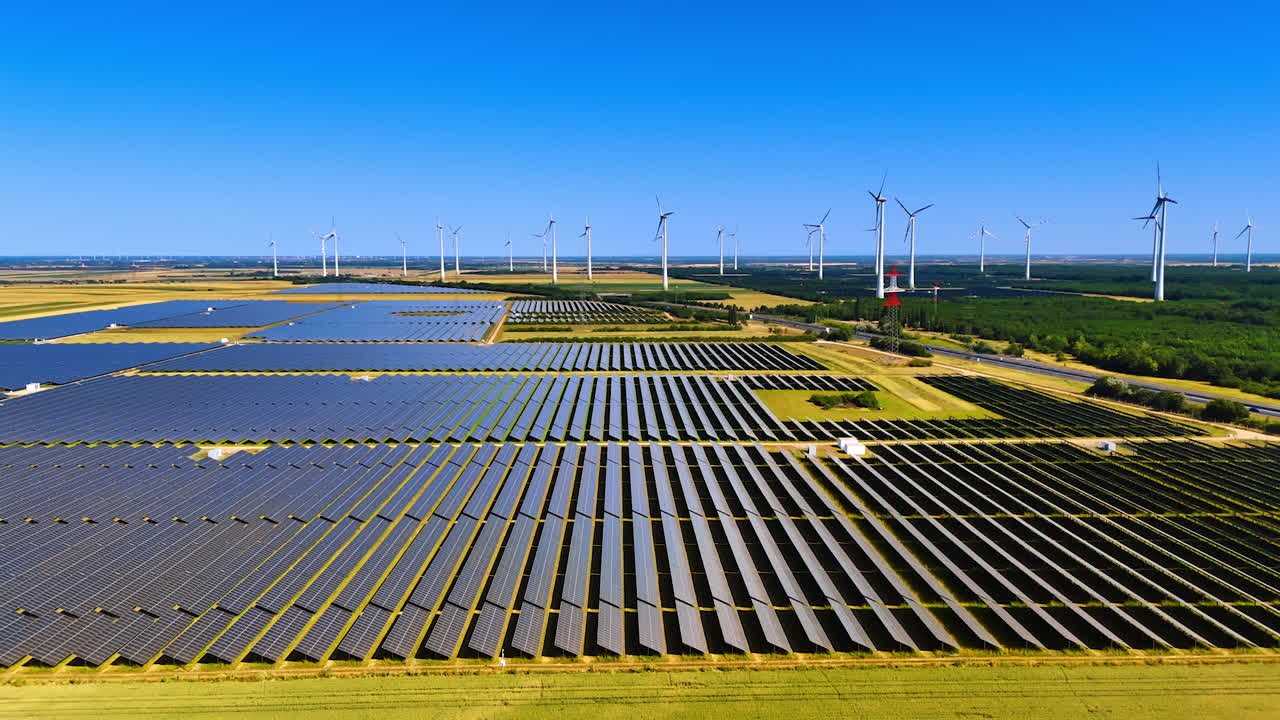 Solar panels turned to the bright sun in the vast fields. Wind mills work at backdrop. Sustainable energy concept. Aerial view