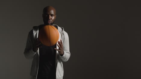 Studio-Portrait-Shot-Of-Male-Basketball-Player-Wearing-Tracksuit-Throwing-Ball-From-Hand-To-Hand-Against-Dark-Background