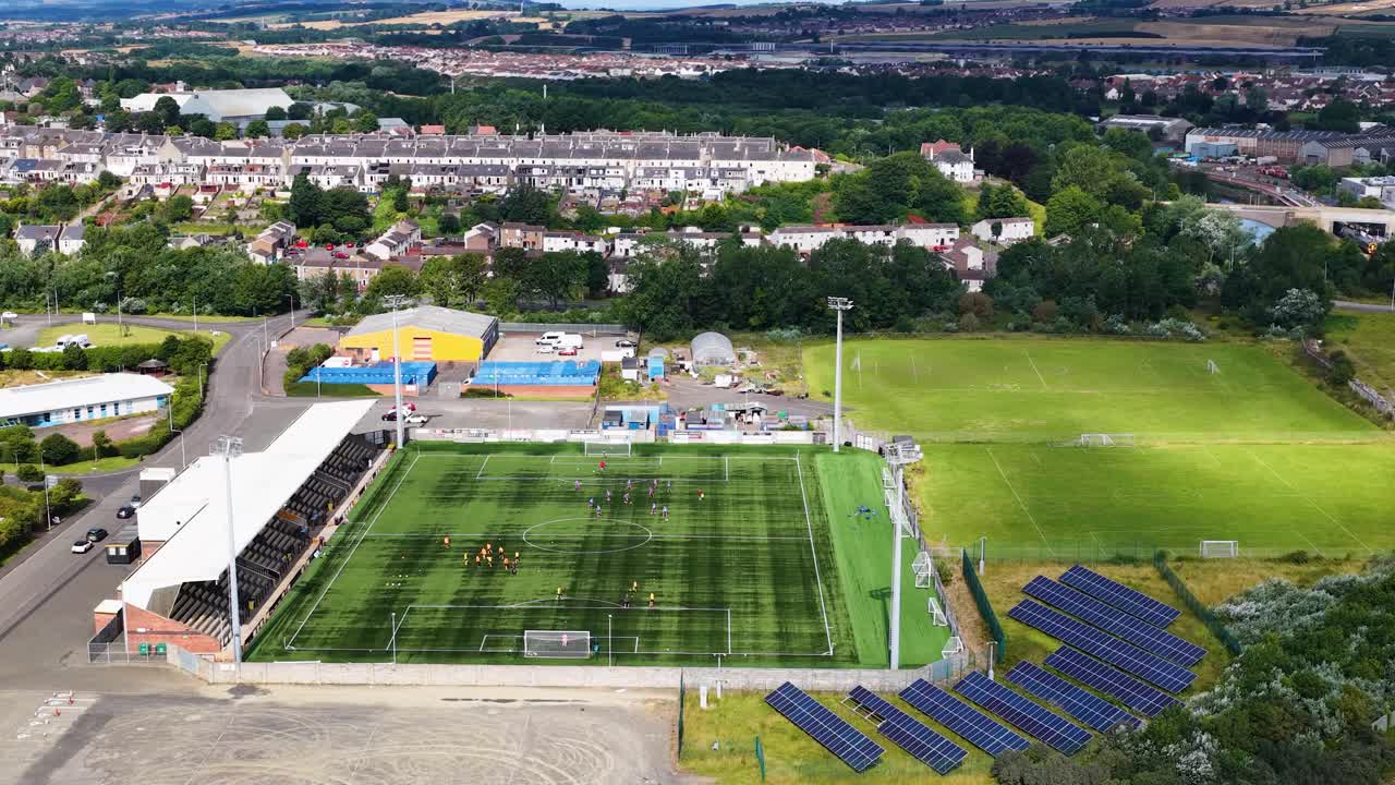 Drone flies over football stadium with solar panels, training fields, and surrounding Fife, Scotland landscape