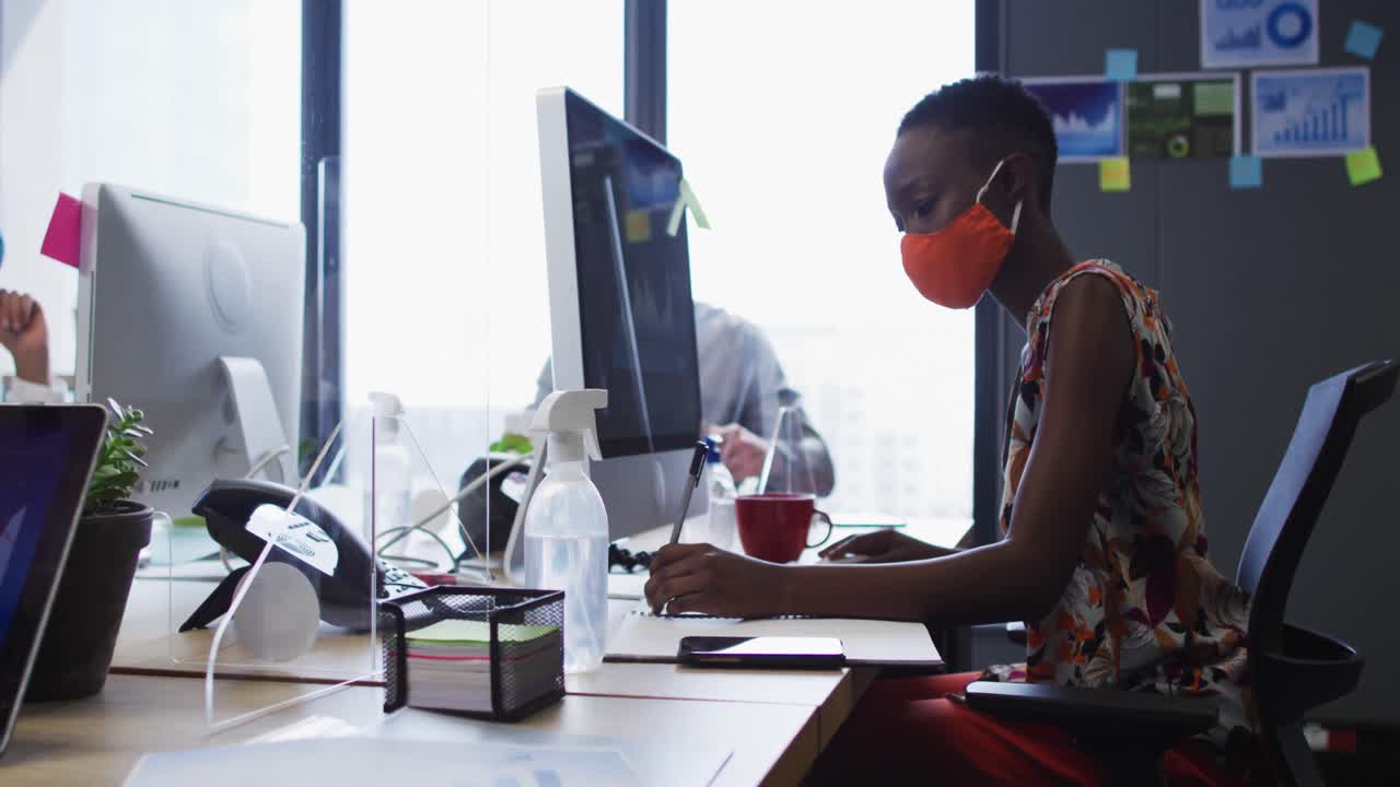 African american woman wearing face mask taking notes while sitting on her desk at modern office
