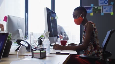 mujer afroamericana con máscara facial tomando notas mientras está sentada en su escritorio en una oficina moderna