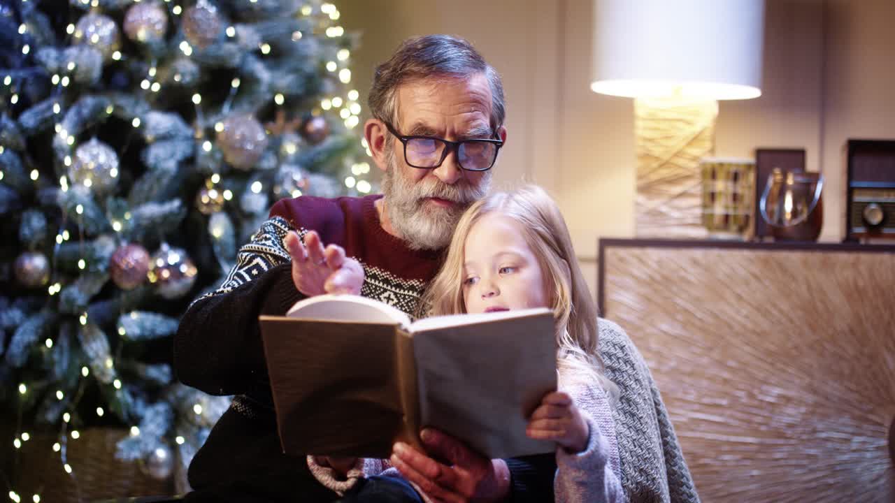 cierra el retrato de un abuelo cariñoso y alegre leyendo un libro a una niña adorable mientras se sienta en casa y pasa la víspera de navidad cerca de un árbol decorado