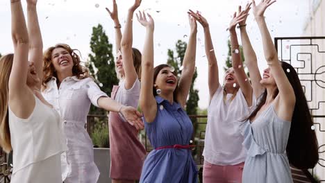 group of young girls dressed in casual partying outdoors on terrace silver confetti in the air at daytime during their carefree hen party. dancing and hugging each other in circle. slow motion