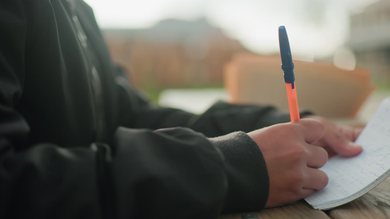 Close up hand view of person writing in notebook with pen on wooden table outdoors while blurred open book in background has pages flipping in wind