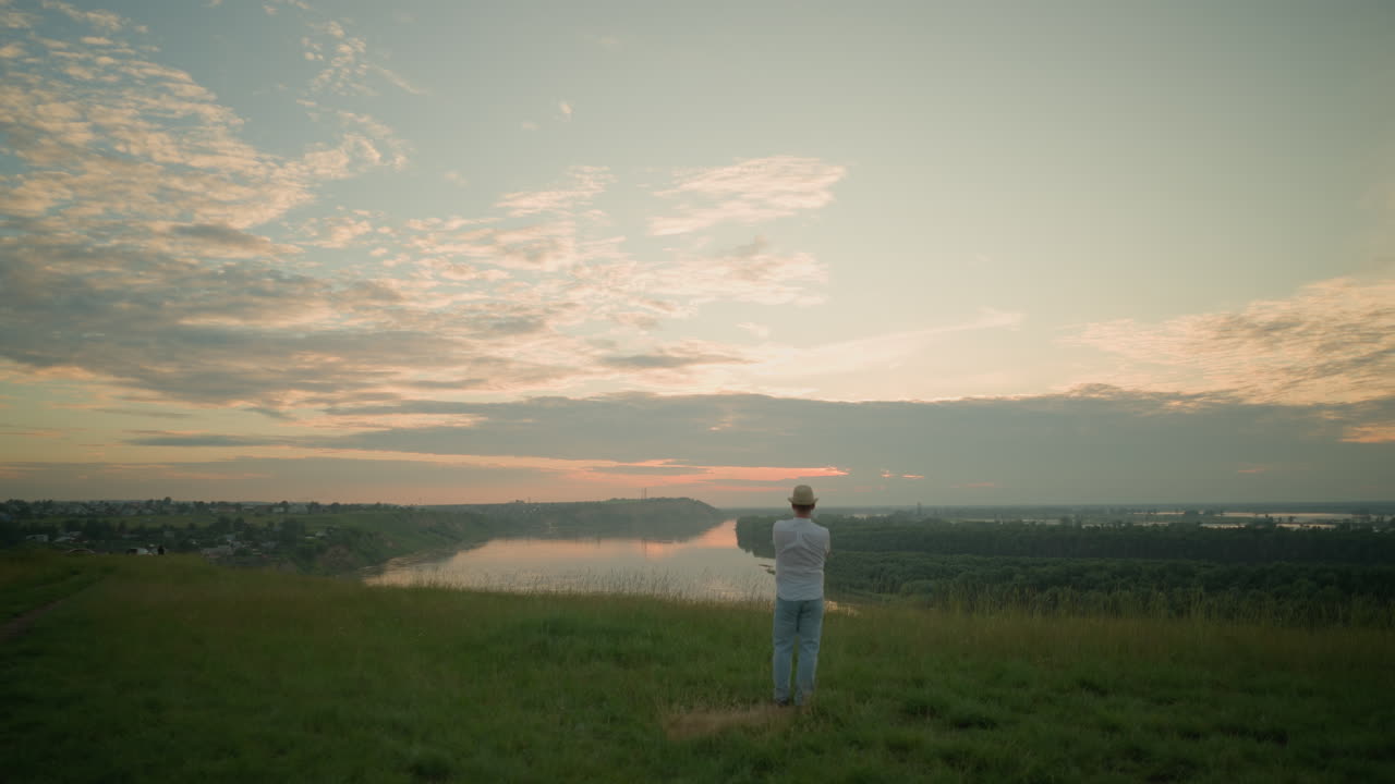 한 신사는 해가 지는 동안 조용한 호수 에 있는 풀에서 상하고 서 있습니다. 색 셔츠, 모자, 청바지를 입고, 그는 해가 지면 평화롭고 반영적인 순간을 포착합니다.