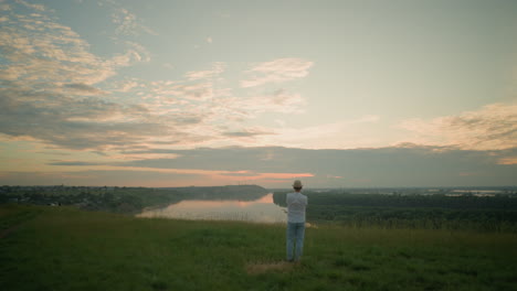 a gentleman stands contemplatively in a grassy field by a tranquil lake at sunset. dressed in a white shirt, hat, and jeans, he captures a peaceful, reflective moment as the sun sets