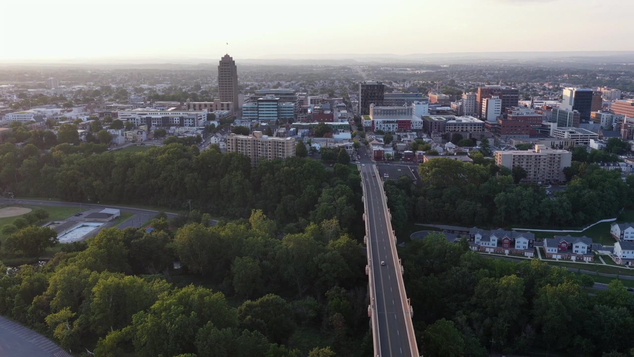 La PPL Tower si erge sopra il centro di Allentown, Pennsylvania; auto attraversano un ponte alberato verso affollati edifici cittadini che brillano caldamente sotto cieli sfocati e dorati di un tramonto serale. Ampia ripresa aerea con drone.