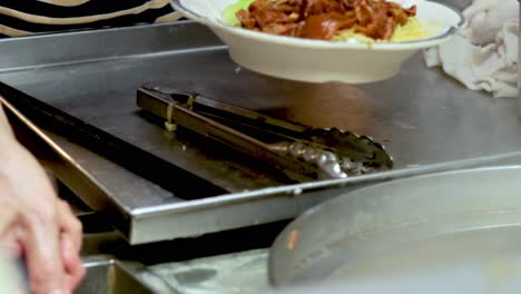 a chef skillfully serves noodles topped with pork and fresh greens in a kitchen setting.