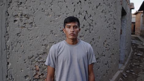 serious young latin male with short dark hair and gray t shirt intensely staring directly at camera, positioned against neutral gray concrete wall during daylight hours