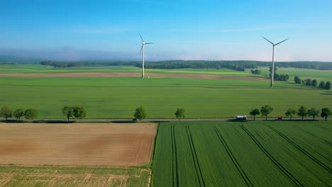 Vehicles-drive-in-front-of-wind-mills,-on-lush-green-fields