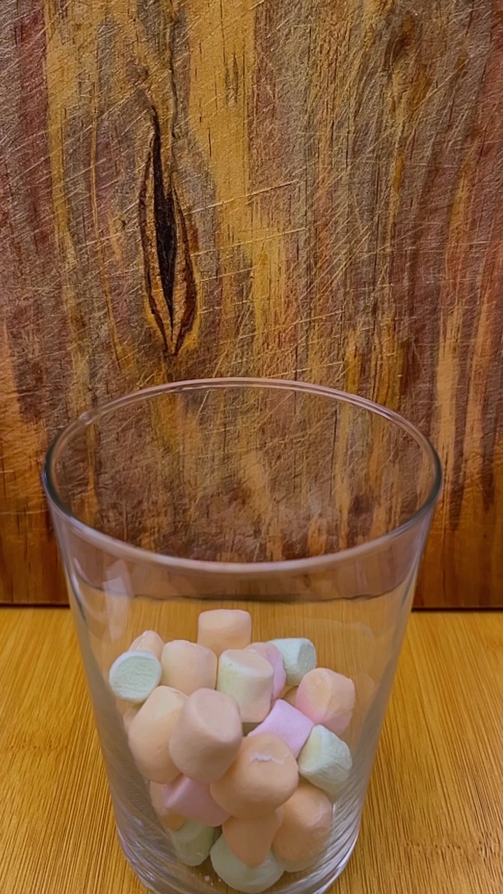 Marshmallow colorati riempiono un bicchiere trasparente posizionato su un tavolo di legno, che mostrano colori pastello tenui e forme arrotondate contro la venatura rustica del legno.