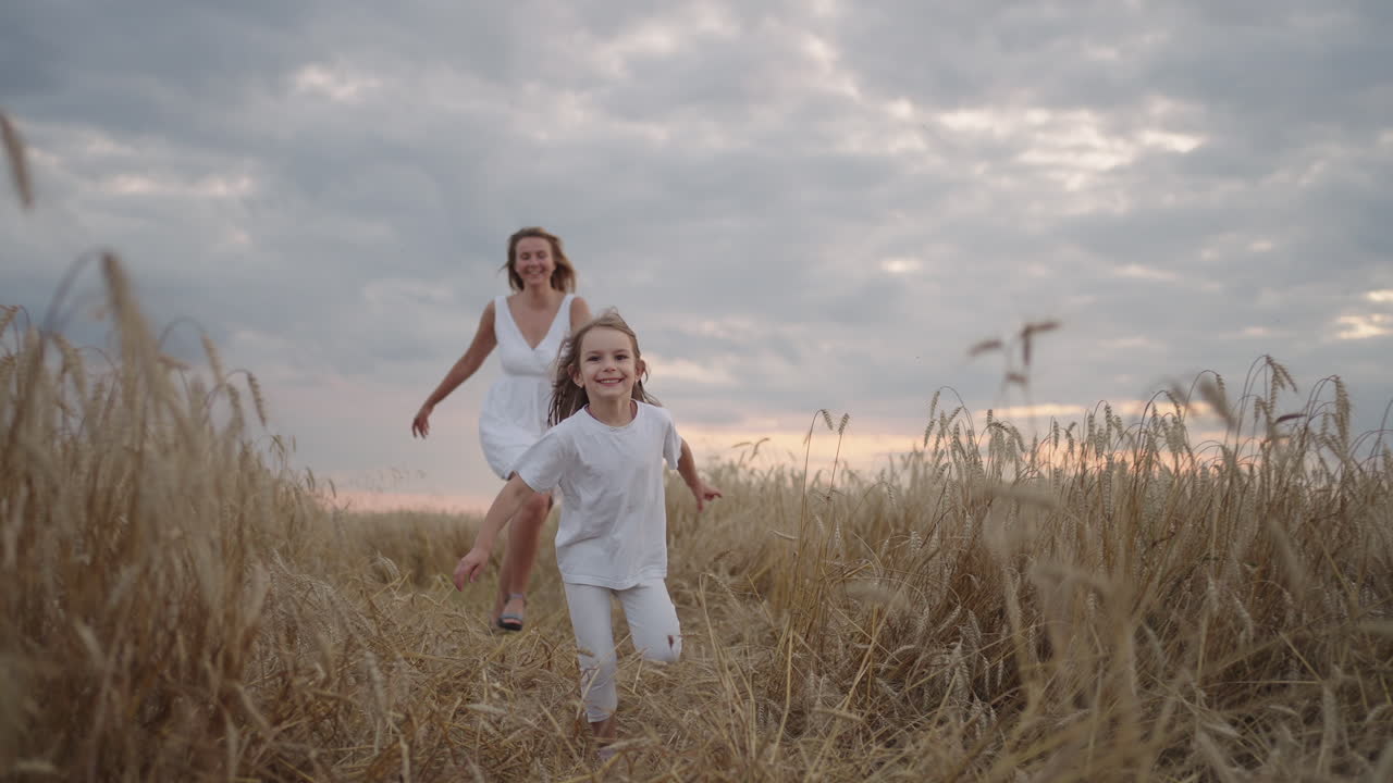Madre e figlia sognano insieme correndo nel campo di grano al tramonto. Concetto di famiglia felice nel campo di grano. Mamma e bambina giocano a rincorrersi. Bambino che corre divertendosi nel prato verde.