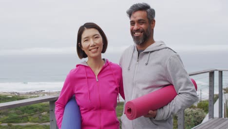 Portrait-of-happy-diverse-couple-holding-yoga-mats-on-balcony