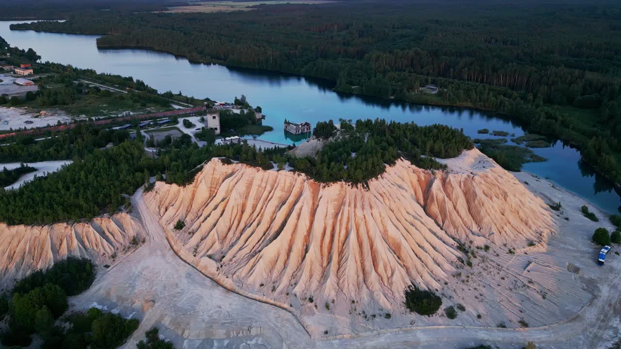 Cinematic drone shot of Rummu Quarry features the towering white ash hill overlooking turquoise water that floods prison ruins and is framed by dense Estonian woodland