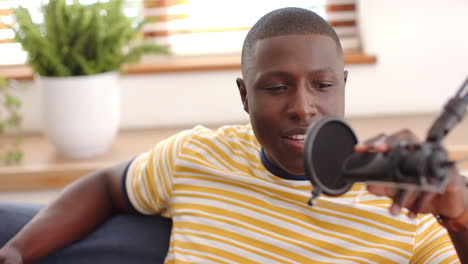 Recording podcast, african american man speaking into microphone while relaxing on couch, at home