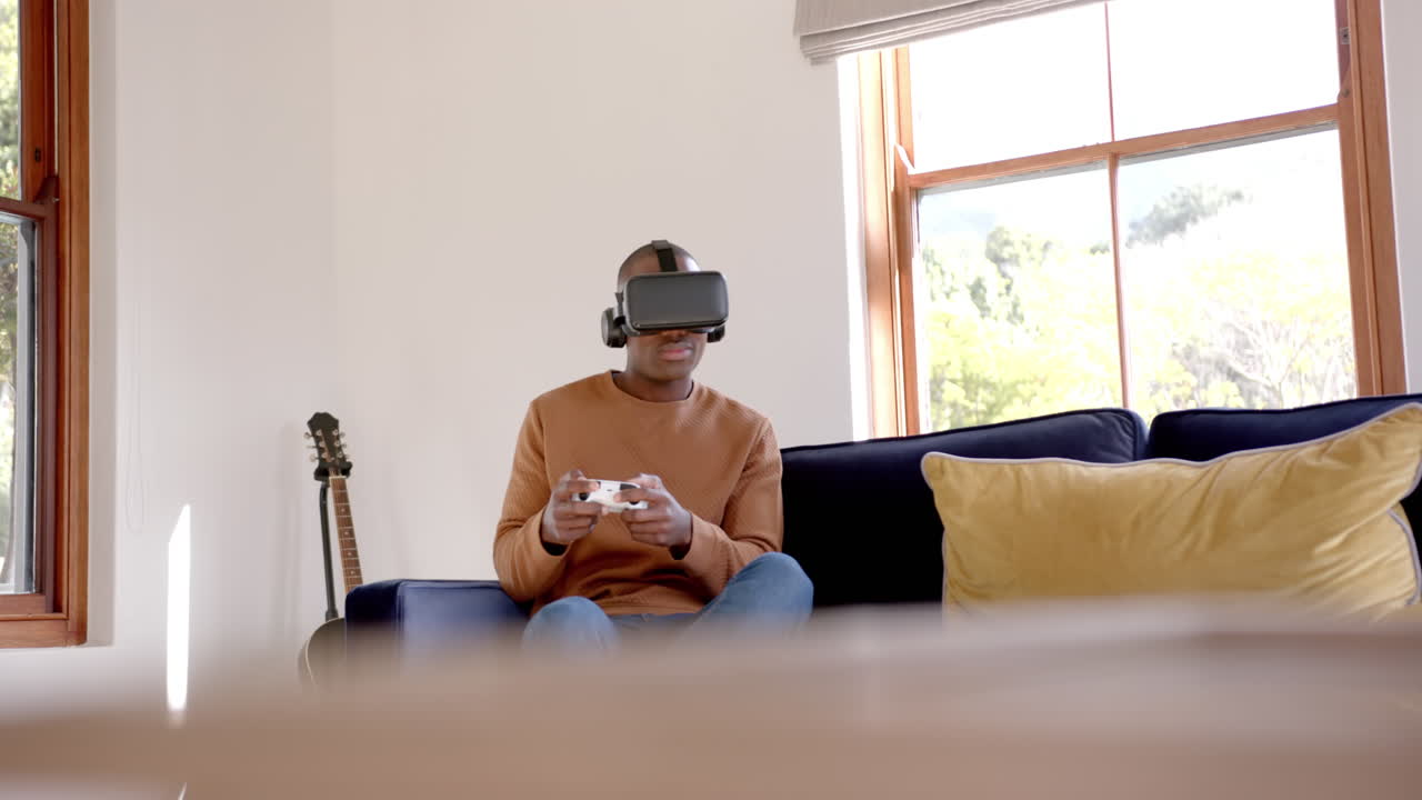 African american man sitting on sofa using vr headset and playing video games at home, slow motion
