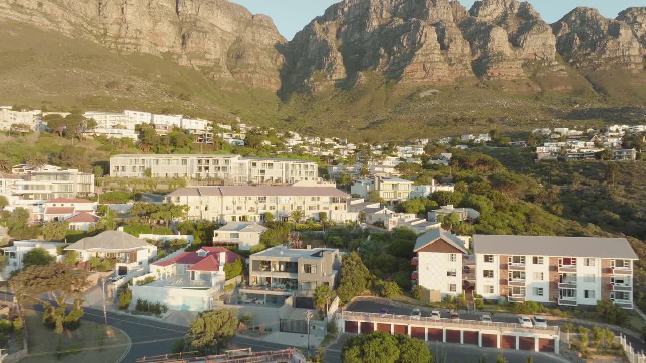 Drone flies fast backwards away from the houses standing on the hillside at Camps Bay beach in Cape Town South Africa - in the background Table Mountain rises illuminated by the sunset