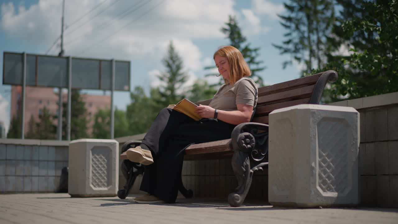 Woman sitting on wooden bench reading book outdoors under clear sky surrounded by green trees and bright sunlight, calm and focused expression showing peaceful learning