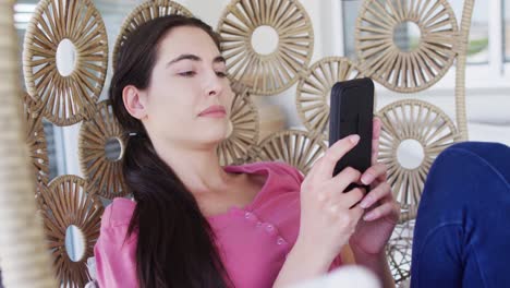 happy caucasian woman sitting on hanging chair on balcony and using smartphone