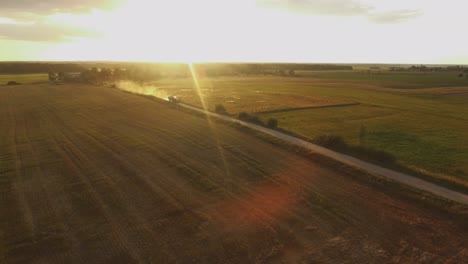 tractor with trailer drives through a rural gravel road leading through agricultural fields on a sunny summer evening