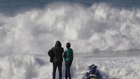 two people watch a jet ski driver from the beach as he rides a foamy wave that throws him high