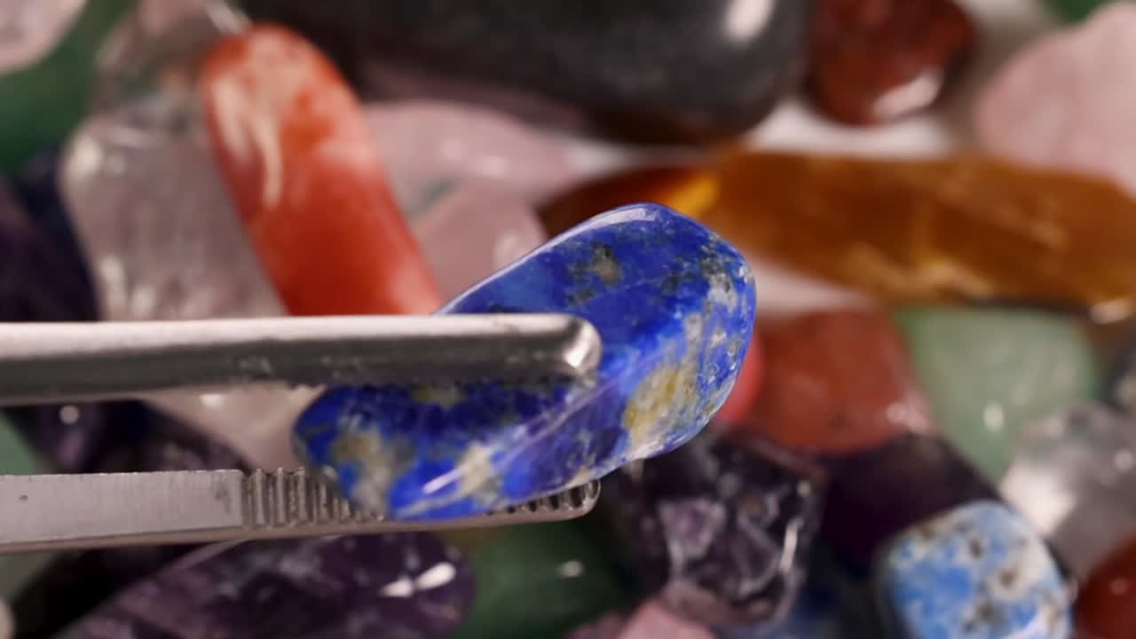 Nahaufnahme einer Pinzette aus Metall, die einen leuchtend blauen Lapislazuli aus bunten Kristallen auswählt