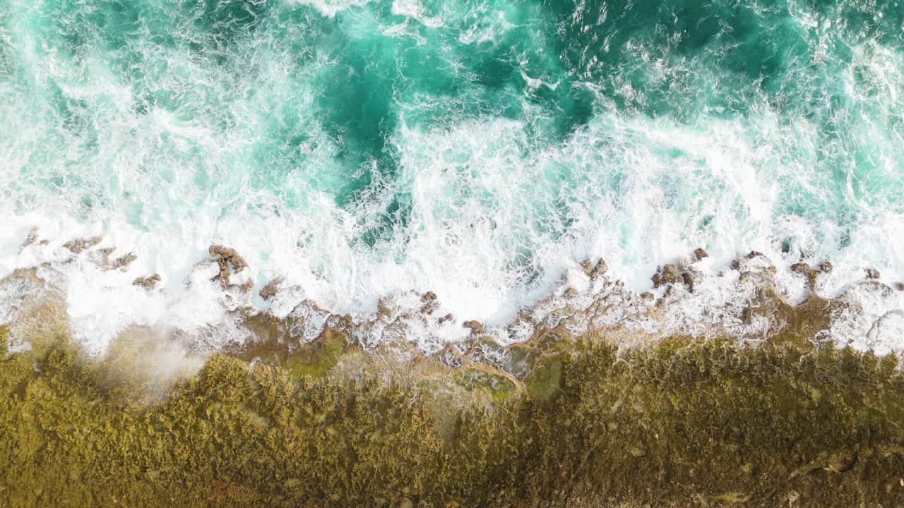Potente acqua oceanica verde e turbolenta si infrange spruzzando nebbia su rocce coperte di alghe in un'isola caraibica, vista drone dall'alto