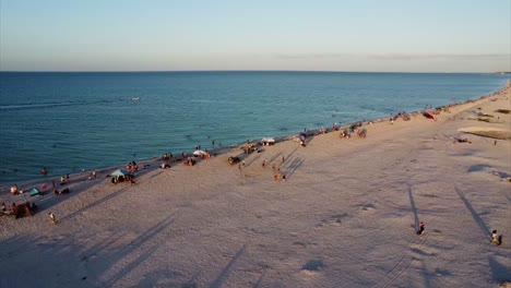 drone shot over beach following fishing boat on blue sea family enjoying white sand during sunset golden hour