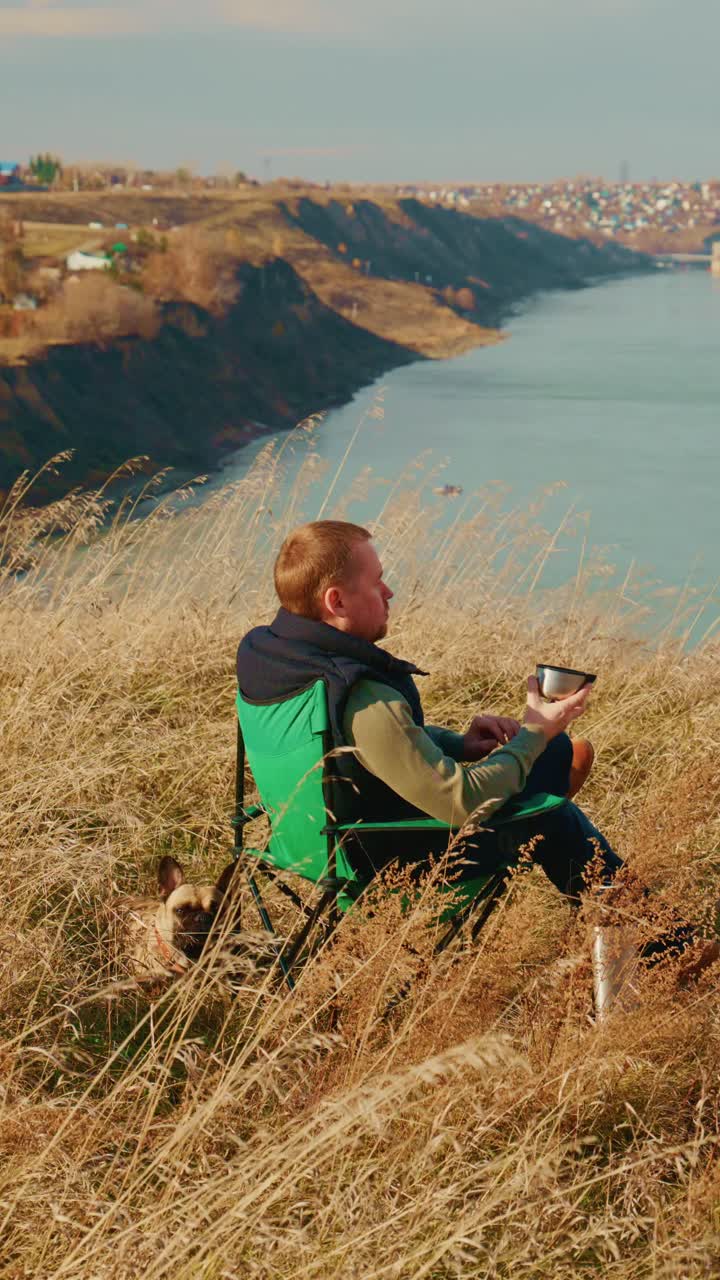 Un momento di tranquillità in riva al fiume, mentre una persona si gode la natura con una vista panoramica, seduta su una sedia accanto al suo fedele animale domestico, immersa nella contemplazione e nel relax tra l'erba rigogliosa e la brezza leggera.