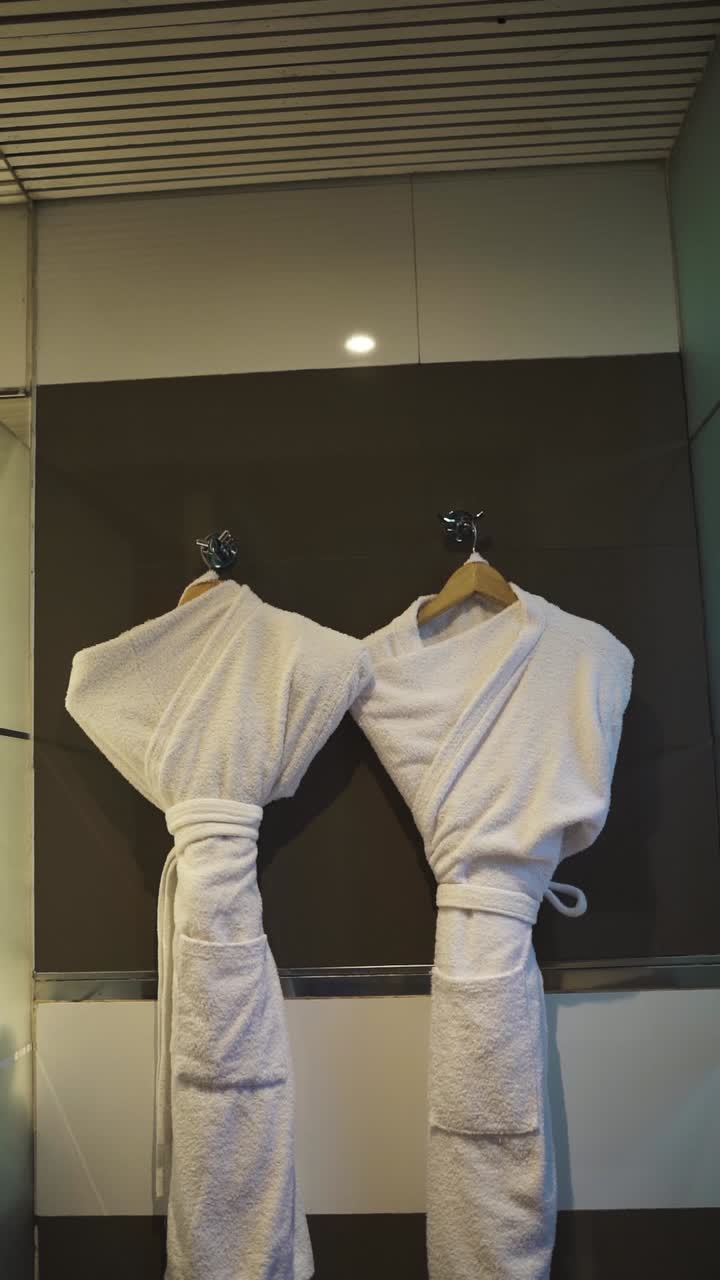 Two white bathrobes hang on wooden hangers against a dark wall in a luxury hotel suite, highlighting the room's modern design and relaxing atmosphere