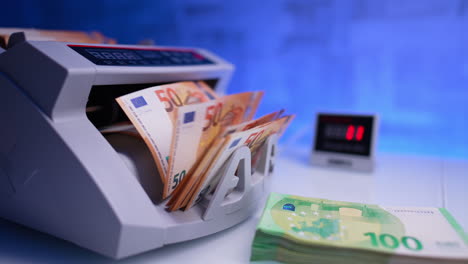 female hand places orange euro bills stack into working cash counting machine. bank equipment quickly processing banknotes. close up. blurred backdrop.