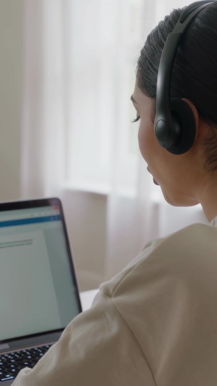 Video vertical: Mujer con auriculares respondiendo a una llamada entrante, leyendo un formulario en una laptop cerca de una ventana