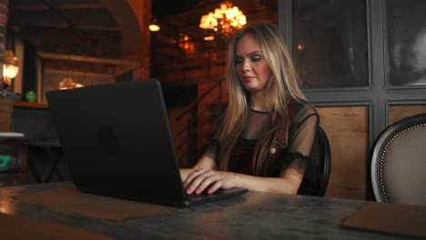 Side-view.-Young-business-woman-sitting-at-table-and-taking-notes-in-notebook.On-table-is-laptop,-smartphone-and-cup-of-coffee.On-computer-screen-graphics-and-charts.-Student-learning-online.-Blogger.