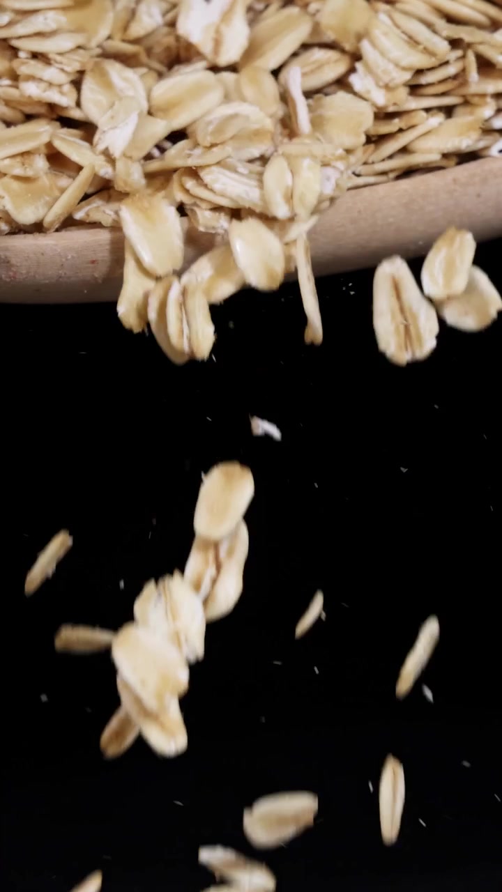 Fiocchi d'avena cadono da un cucchiaio al rallentatore, su uno sfondo scuro, evidenziandone la consistenza e il movimento.