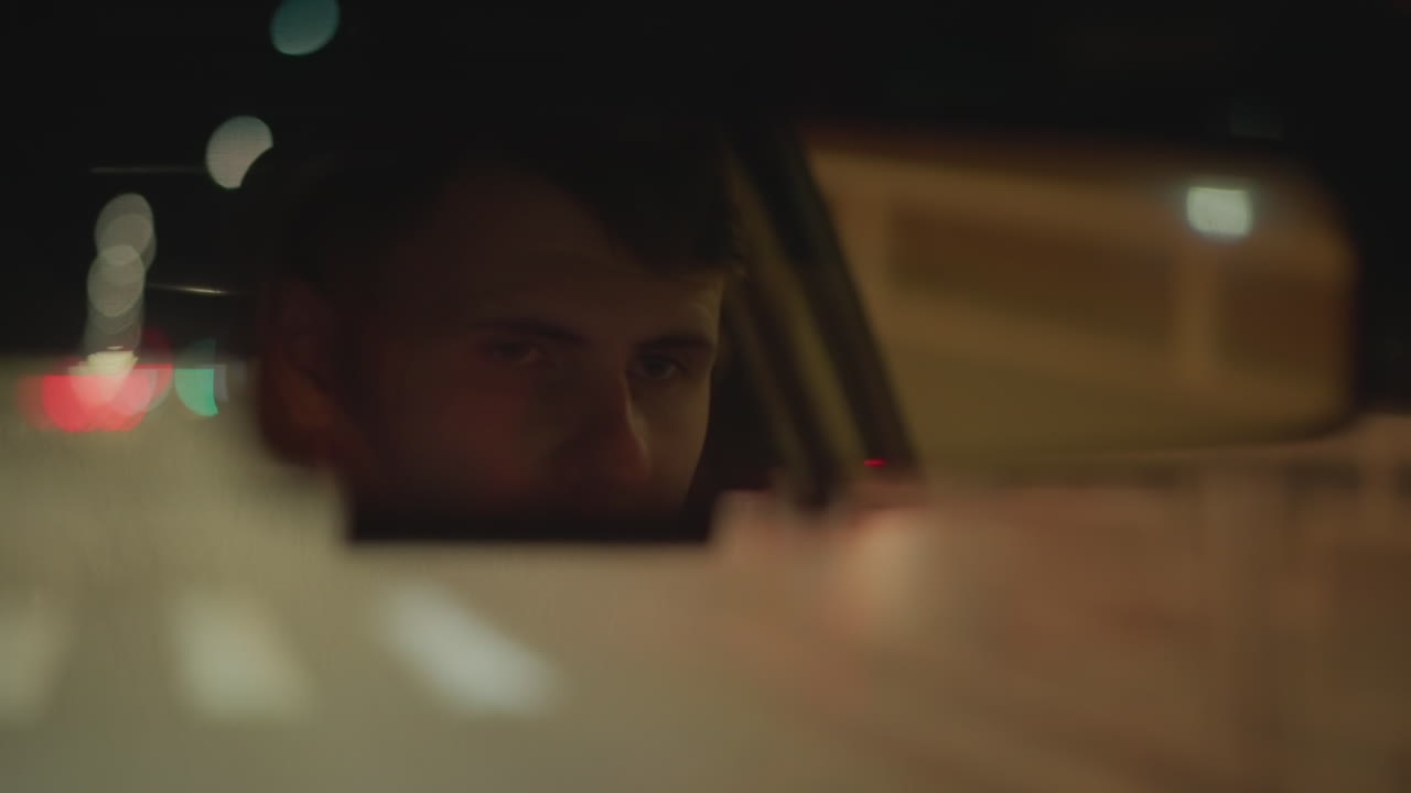 autista caucasico nello specchietto di notte, sguardo pensieroso e bokeh tenue della città, bagliore fioco del cruscotto, riflessi del traffico suburbano, fotogrammi cinematografici in stile noir suggeriscono un pendolare che fa il turno di notte, un tassista stanco