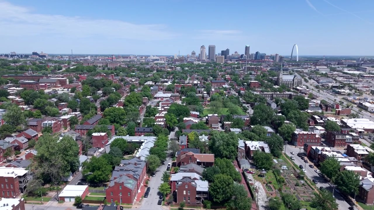 Blocchi residenziali in mattoni e strade alberate a St. Louis scorrono via mentre l'inquadratura vola verso lo skyline del centro e il Gateway Arch in una giornata limpida