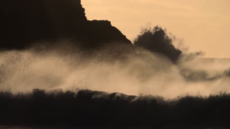 massive waves break against a rocky shore in golden light in slow motion