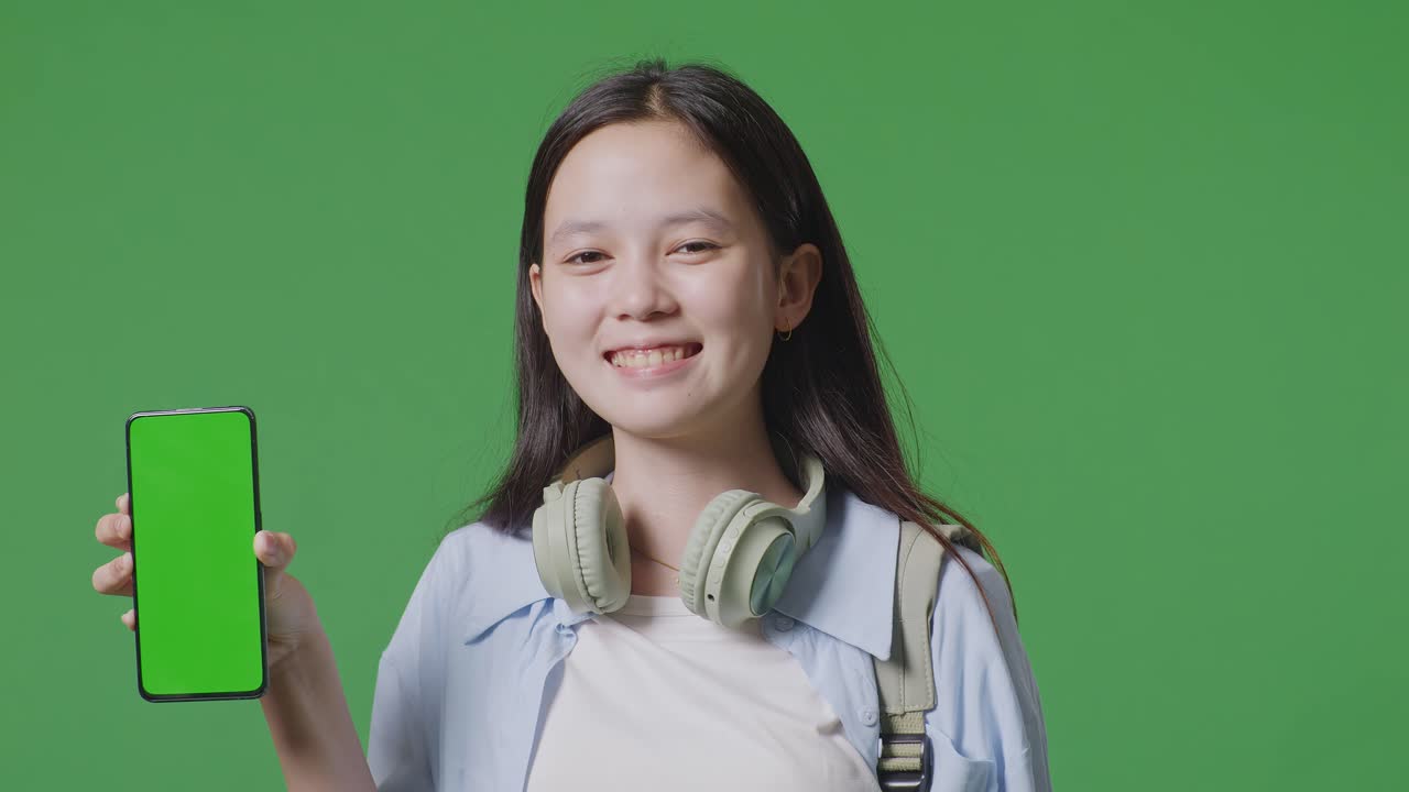 녹색 스크린 배경 스튜디오에 서있는 동안 카메라에 녹색 화면 스마트폰을 보여주고 미소 짓는 배을 가진 아시아 십대 소녀 학생의 클로즈업