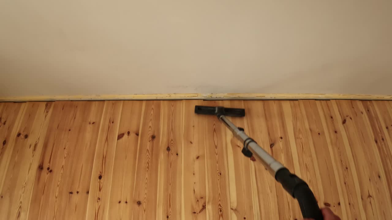 Wide-angle shot of a worker vacuuming polished wooden flooring near a wall, showing professional cleaning technique and smooth tool movement across bright natural pine